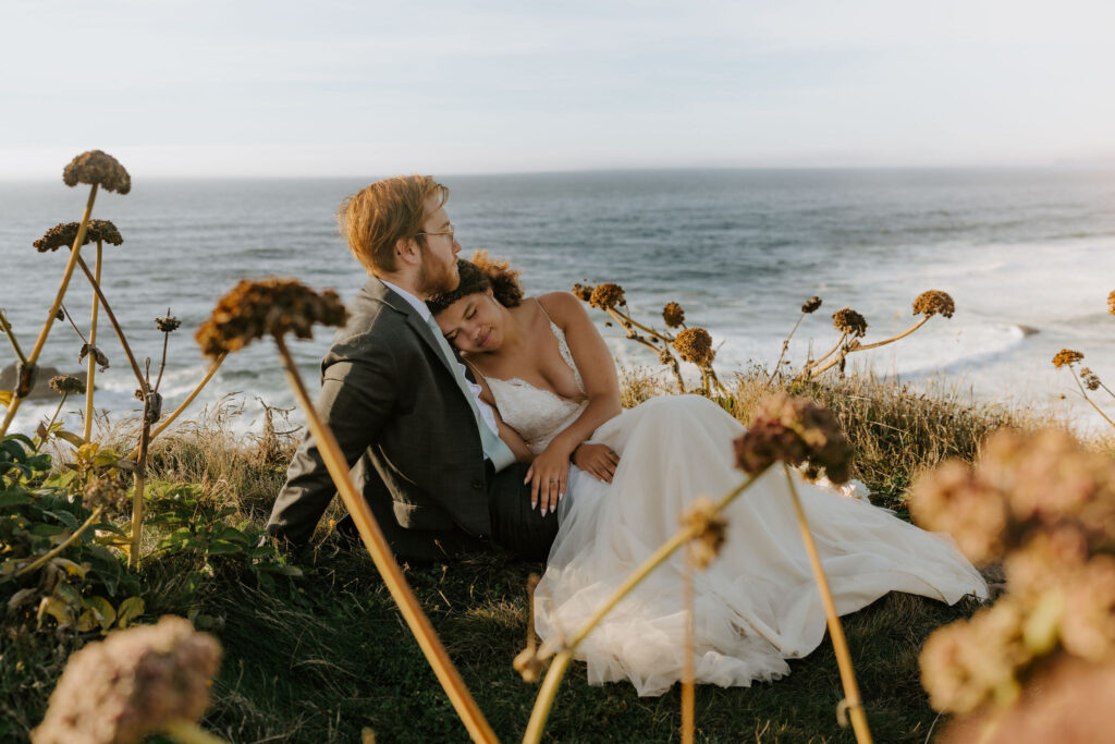 cuddly elopement couple sitting in the grass with ocean views in the backdrop