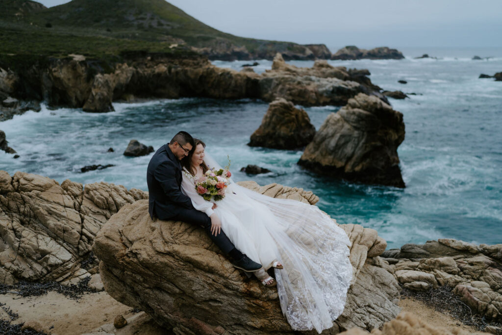bride and groom sitting on a large rock at the coast in Big Sur