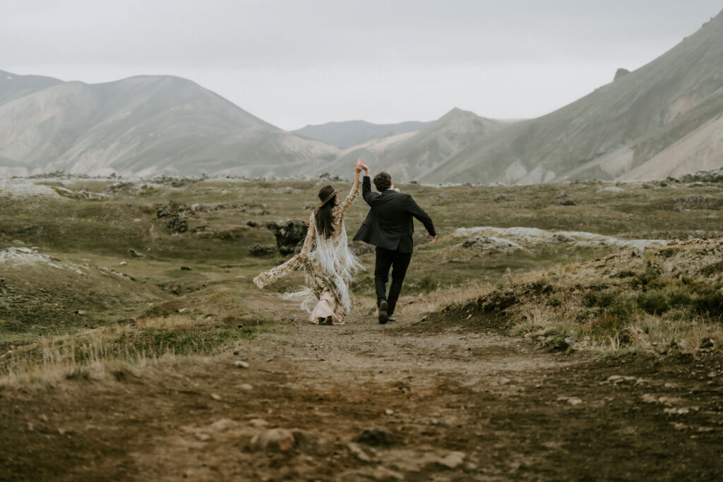 Couple running through the rugged Icelandic Highlands, laughing and windblown, with the bride’s embroidered fringe dress trailing behind her.