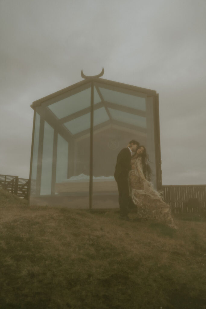 Couple embracing in front of a sleek glass lodge in the Icelandic Highlands, sharing a quiet moment on the grassy slope.