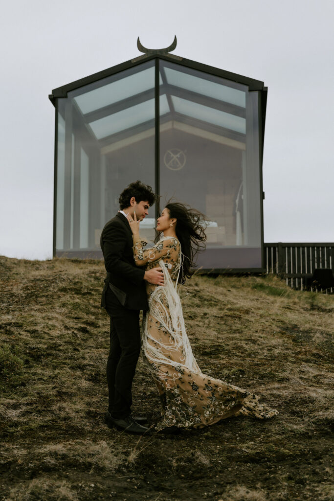 Couple embracing in front of a sleek glass lodge in the Icelandic Highlands, sharing a quiet moment on the grassy slope.
