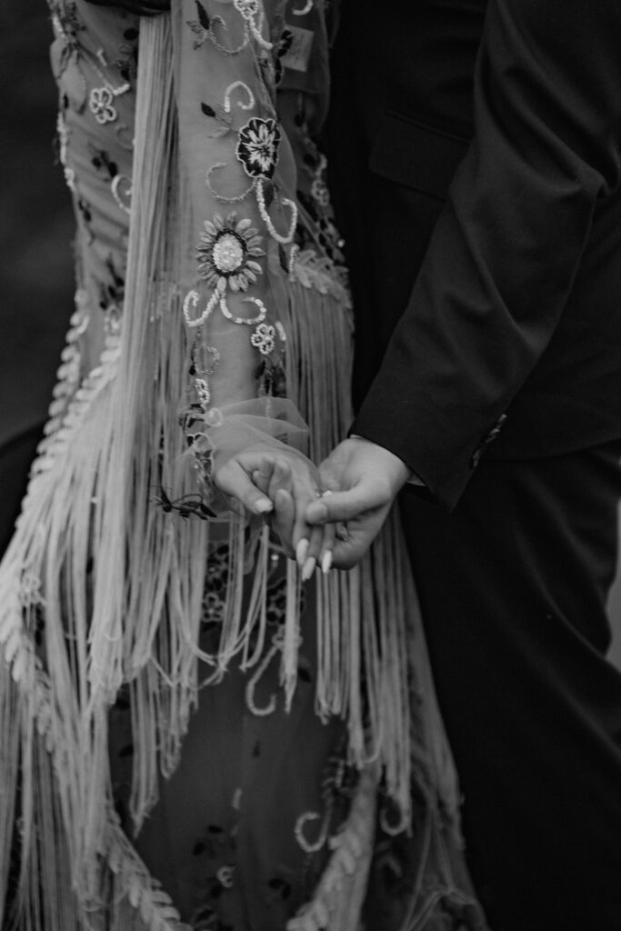 close up of bride and groom holding hands, bride wearing a boho western inspired dress with fringe sleeves