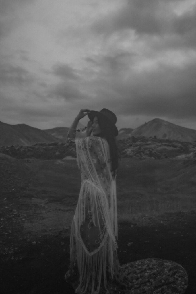 Bride alone in black-and-white, holding her hat in a windswept landscape, with moody skies at Landmannalaugar, Iceland