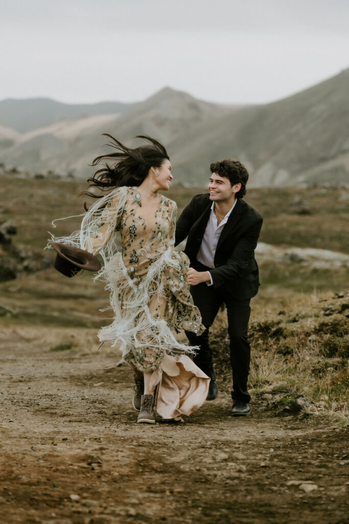 Couple running through the rugged Icelandic Highlands, laughing and windblown, with the bride’s embroidered fringe dress trailing behind her.